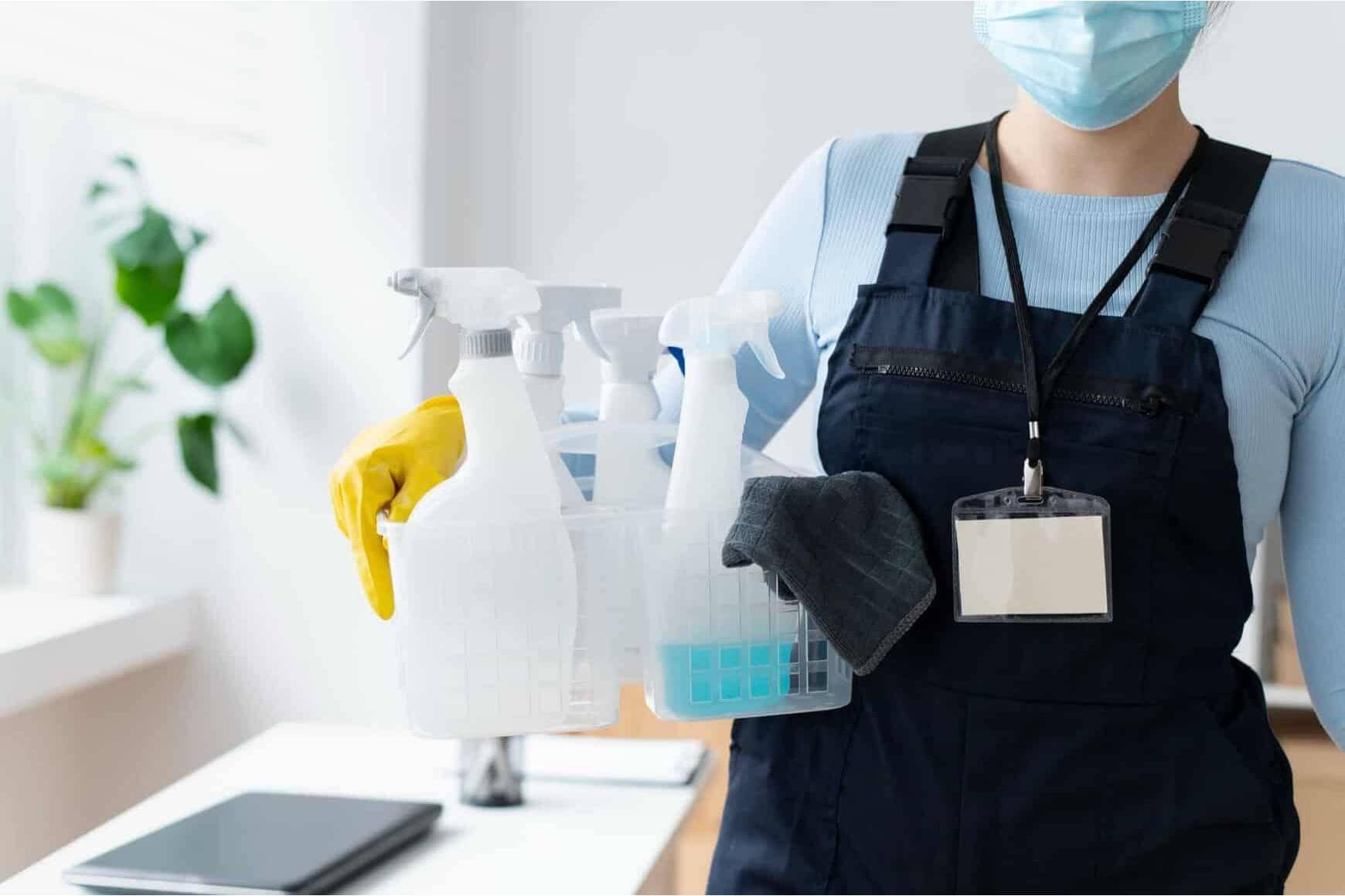 femeie care ține în mână mai multe produse de curățenie, bluză albastru deschis, mănușă galbenă, salopetă